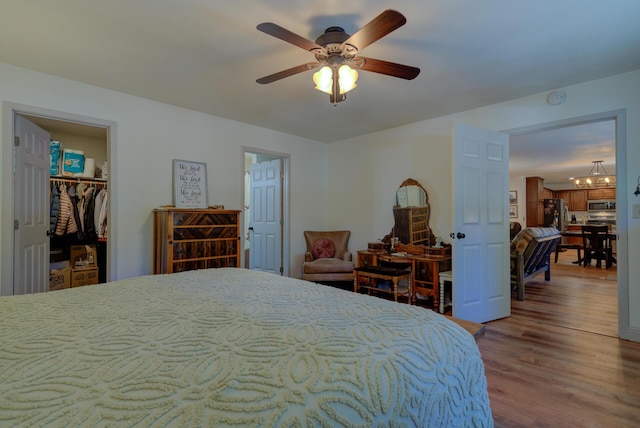 bedroom featuring a walk in closet, freestanding refrigerator, wood finished floors, and ceiling fan with notable chandelier