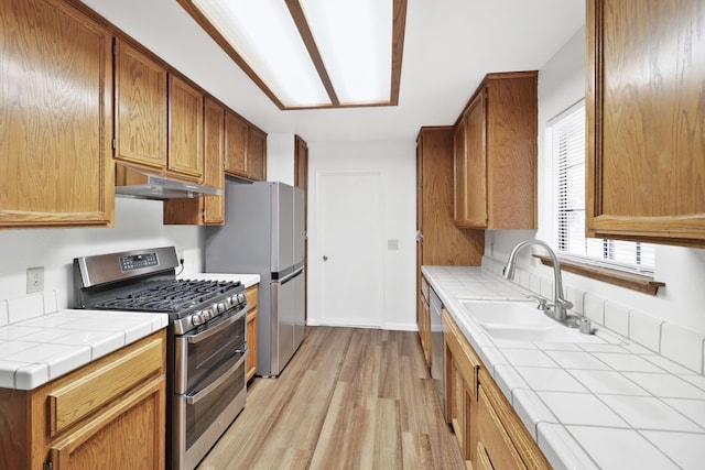 kitchen featuring stainless steel appliances, under cabinet range hood, a sink, tile counters, and light wood-style flooring