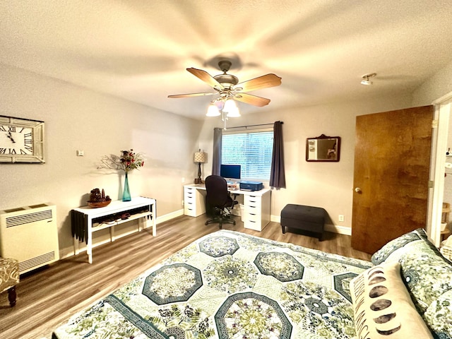 bedroom with heating unit, wood finished floors, baseboards, a textured ceiling, and a ceiling fan