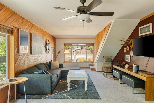 living room featuring carpet flooring, wooden walls, a ceiling fan, and stairway