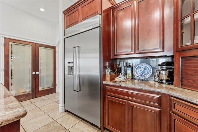 kitchen with light stone countertops, glass insert cabinets, light tile patterned floors, built in fridge, and backsplash