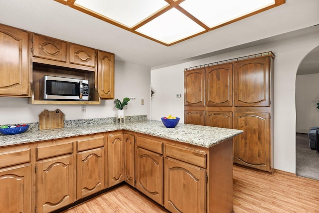 kitchen featuring stainless steel microwave, a peninsula, arched walkways, light wood-type flooring, and brown cabinetry