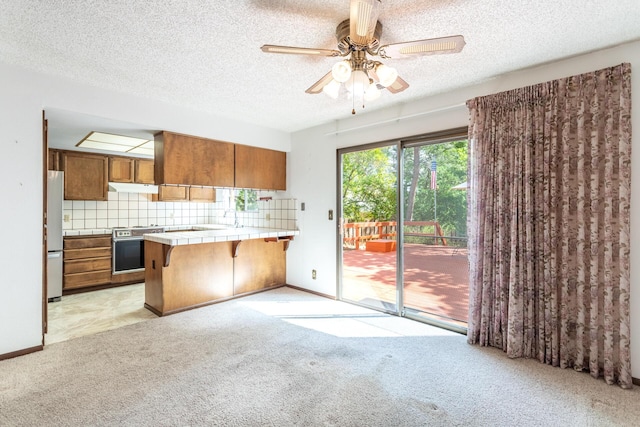 kitchen featuring tile counters, brown cabinets, light colored carpet, a peninsula, and wall oven