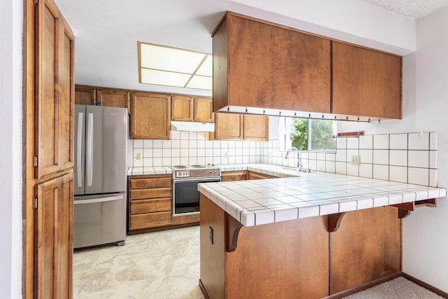 kitchen featuring appliances with stainless steel finishes, a peninsula, a sink, brown cabinets, and decorative backsplash