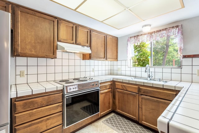 kitchen featuring under cabinet range hood, stainless steel range with electric stovetop, tile countertops, a sink, and decorative backsplash