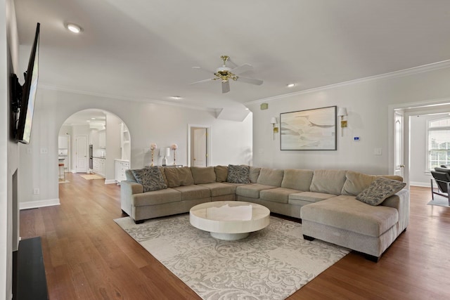 living room with arched walkways, wood finished floors, a ceiling fan, baseboards, and crown molding