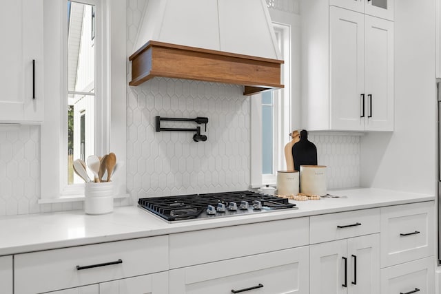 kitchen with custom range hood, glass insert cabinets, stainless steel gas stovetop, white cabinetry, and backsplash