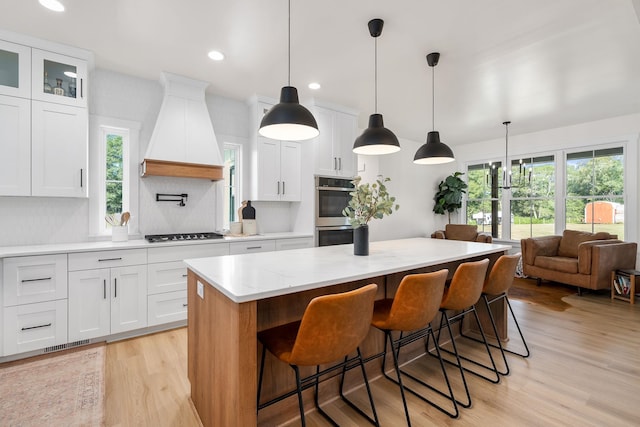 kitchen featuring double oven, premium range hood, a kitchen island, white cabinetry, and light wood-type flooring