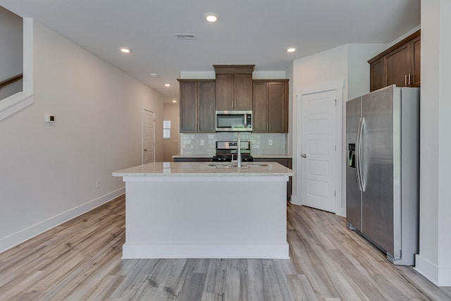 kitchen featuring light wood-type flooring, light stone counters, an island with sink, sink, and appliances with stainless steel finishes