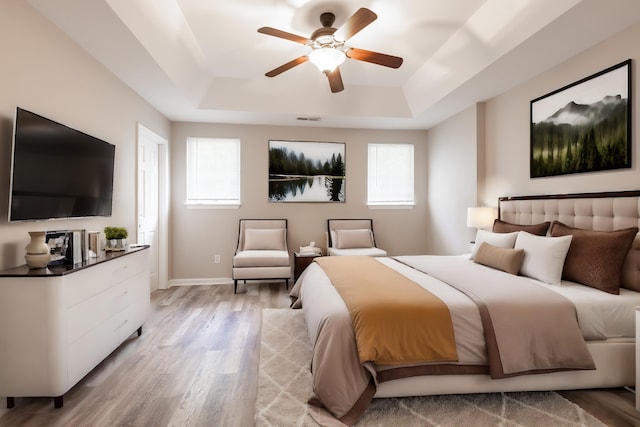 bedroom featuring light wood-type flooring, multiple windows, a raised ceiling, and ceiling fan