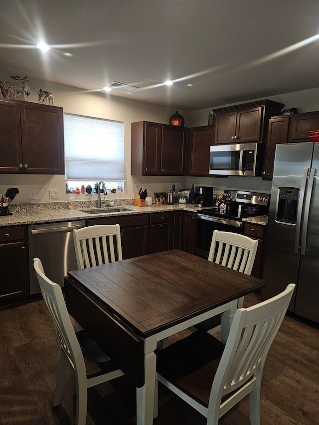 kitchen with appliances with stainless steel finishes, dark brown cabinets, light stone countertops, sink, and dark wood-type flooring