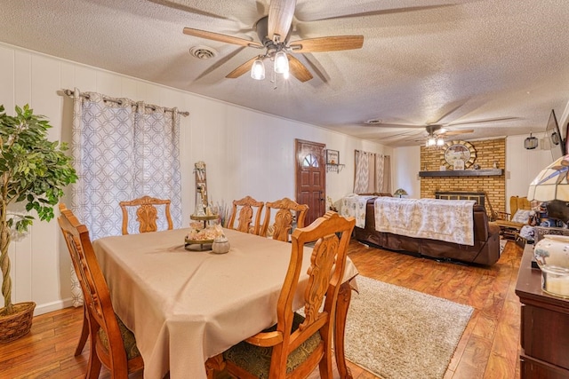 dining room with a brick fireplace, ceiling fan, wood-type flooring, and a textured ceiling