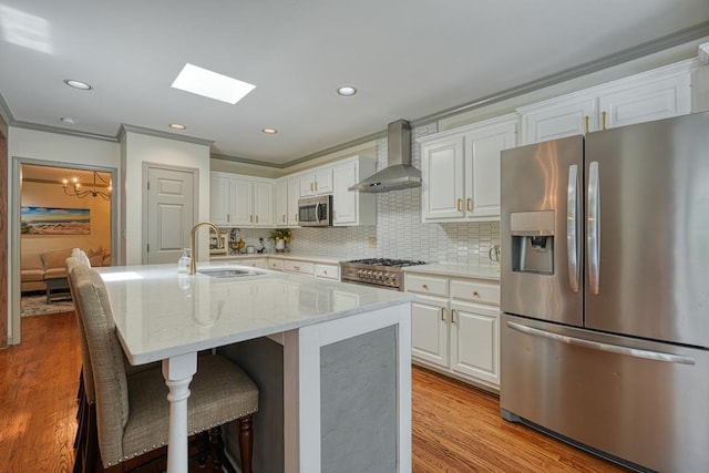 kitchen with light wood-type flooring, stainless steel appliances, a kitchen island with sink, wall chimney range hood, and white cabinets