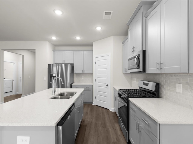 kitchen with appliances with stainless steel finishes, washer / dryer, sink, a kitchen island with sink, and dark wood-type flooring