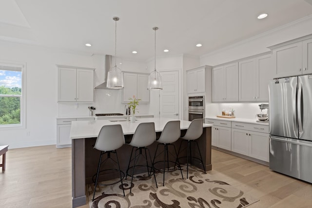 kitchen featuring appliances with stainless steel finishes, light wood-type flooring, wall chimney range hood, a center island with sink, and hanging light fixtures