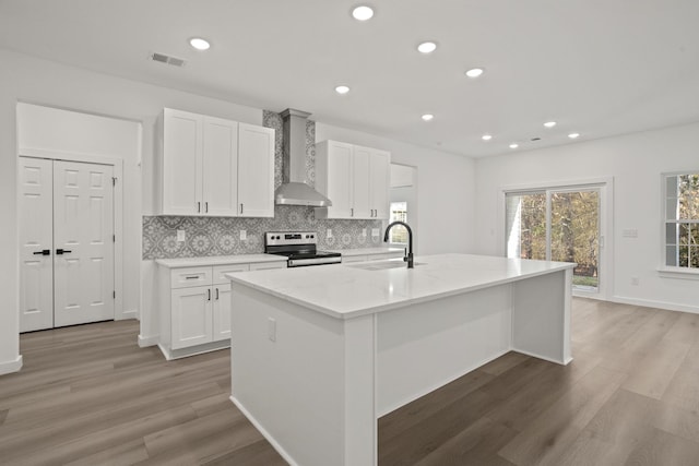 kitchen featuring white cabinets, stainless steel electric range oven, wall chimney exhaust hood, and an island with sink