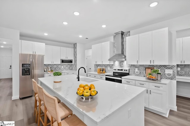 kitchen with sink, wall chimney exhaust hood, stainless steel appliances, a center island with sink, and light wood-type flooring