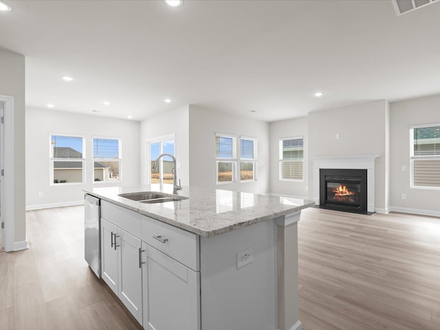 kitchen with an island with sink, sink, stainless steel dishwasher, light stone counters, and light hardwood / wood-style flooring