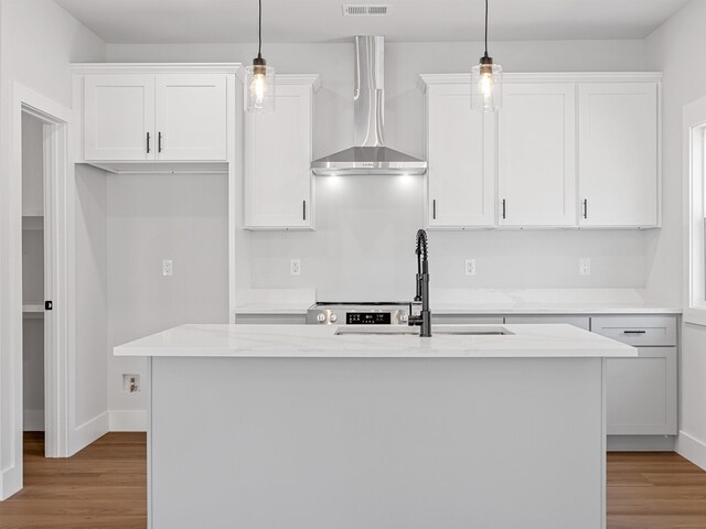 kitchen featuring wall chimney exhaust hood, white cabinetry, an island with sink, and hanging light fixtures