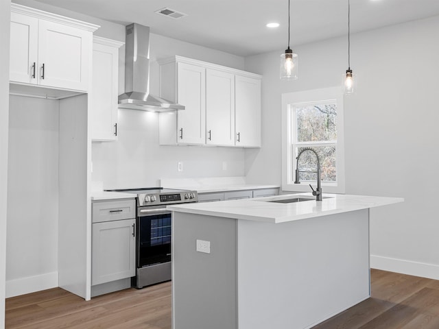 kitchen featuring stainless steel electric range oven, a center island with sink, white cabinets, and wall chimney range hood