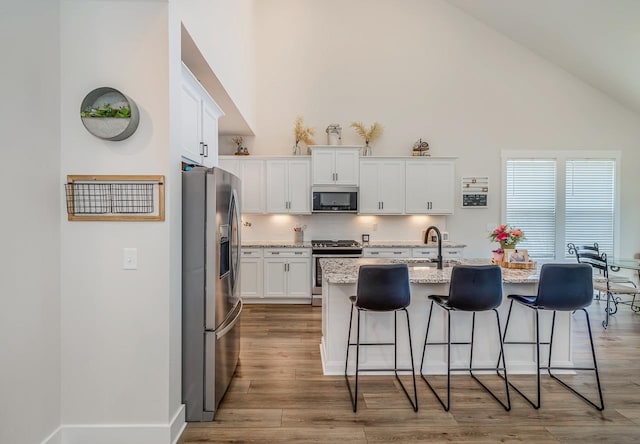 kitchen featuring an island with sink, stainless steel appliances, white cabinetry, and light stone counters