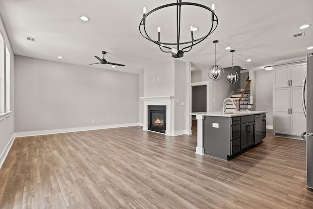 kitchen with light stone counters, decorative light fixtures, an island with sink, light wood-type flooring, and ceiling fan with notable chandelier