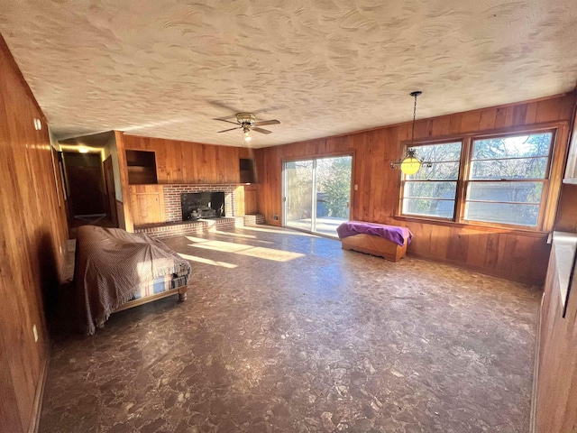 unfurnished living room with ceiling fan, a brick fireplace, wooden walls, and a textured ceiling