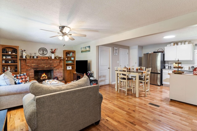 living room with a textured ceiling, a brick fireplace, light hardwood / wood-style floors, and ceiling fan