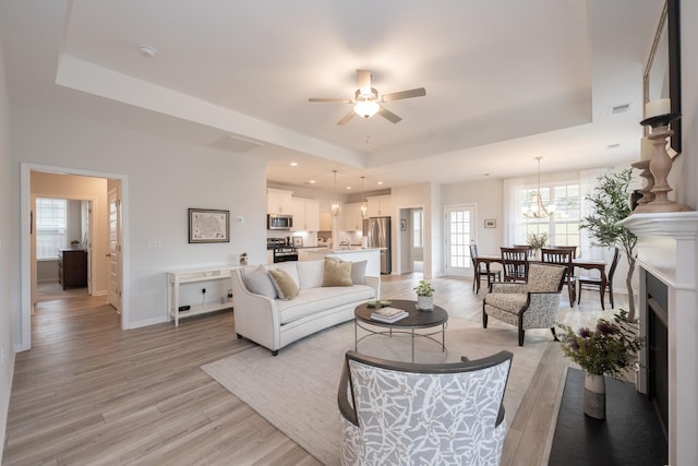 living room featuring light wood-type flooring, ceiling fan with notable chandelier, and a tray ceiling