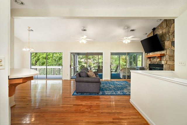 living room featuring hardwood / wood-style flooring, lofted ceiling with beams, a stone fireplace, and ceiling fan with notable chandelier