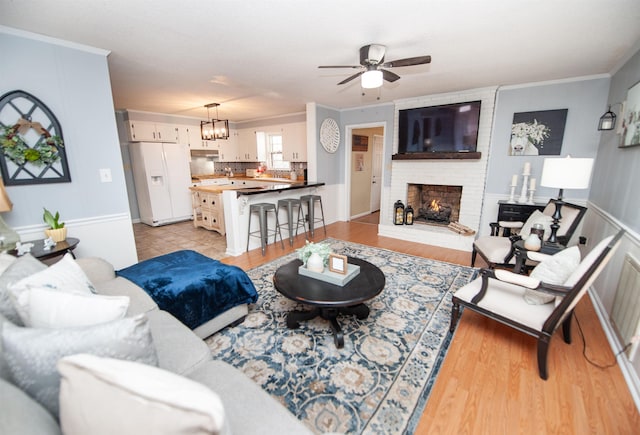 living room featuring a brick fireplace, ceiling fan with notable chandelier, crown molding, and light wood-type flooring