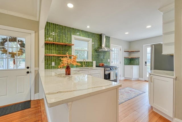 kitchen with stainless steel gas stove, white cabinetry, kitchen peninsula, light stone countertops, and wall chimney range hood