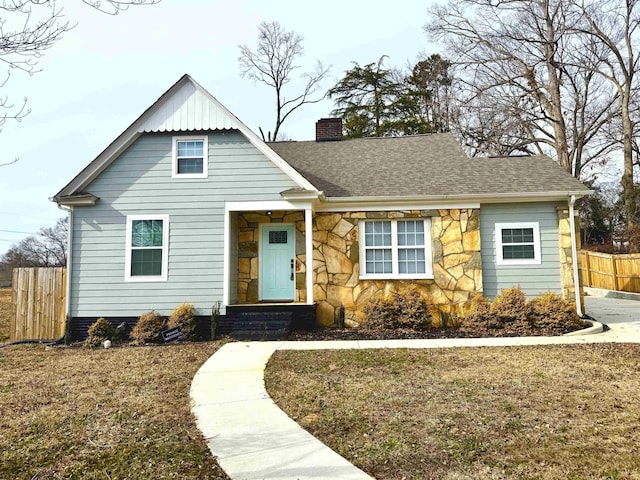 view of front of house featuring stone siding, a shingled roof, a chimney, and fence