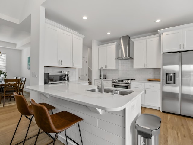 kitchen featuring appliances with stainless steel finishes, white cabinetry, a kitchen breakfast bar, wall chimney exhaust hood, and light wood-type flooring