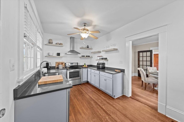 kitchen with sink, stainless steel range with electric stovetop, ceiling fan, wall chimney range hood, and light hardwood / wood-style flooring