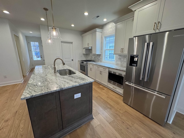 kitchen with an island with sink, sink, hanging light fixtures, stainless steel appliances, and light stone countertops