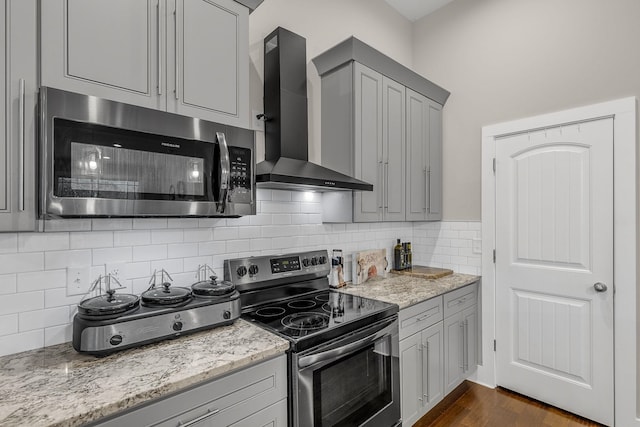 kitchen featuring stainless steel appliances, light stone counters, wall chimney range hood, decorative backsplash, and gray cabinets