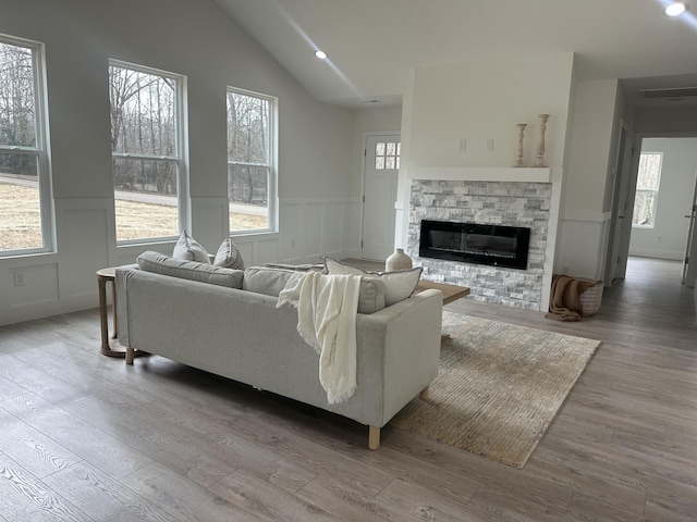 living area featuring a stone fireplace, a wainscoted wall, visible vents, vaulted ceiling, and light wood-type flooring