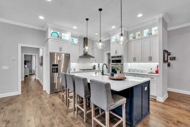 kitchen with white cabinetry, appliances with stainless steel finishes, wall chimney exhaust hood, an island with sink, and glass insert cabinets