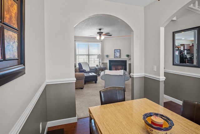 dining space with a warm lit fireplace, baseboards, dark wood-style floors, ceiling fan, and a textured ceiling