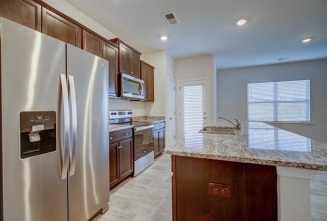 kitchen with light stone counters, light wood-style flooring, a sink, appliances with stainless steel finishes, and an island with sink