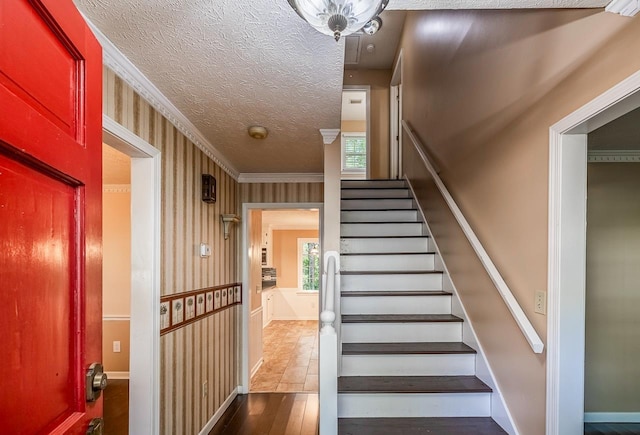 staircase with ornamental molding, a textured ceiling, wood finished floors, baseboards, and wallpapered walls