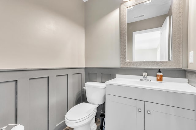 half bath with visible vents, toilet, a wainscoted wall, vanity, and a decorative wall