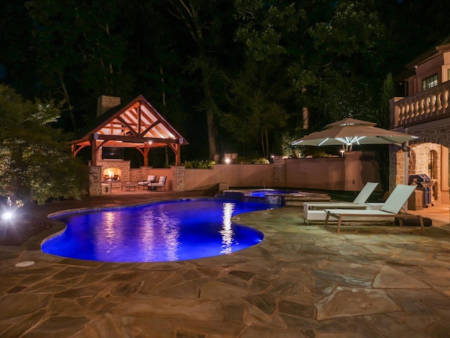 pool at night with a fireplace, a patio, a pool with connected hot tub, grilling area, and a gazebo