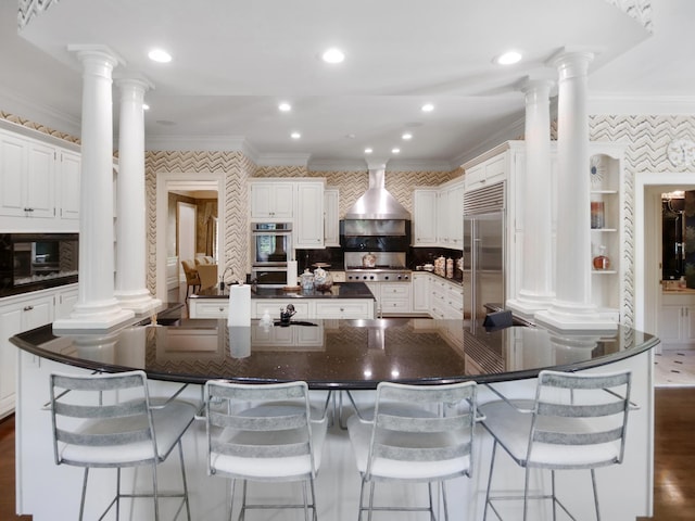 kitchen with appliances with stainless steel finishes, wall chimney range hood, ornate columns, wallpapered walls, and crown molding