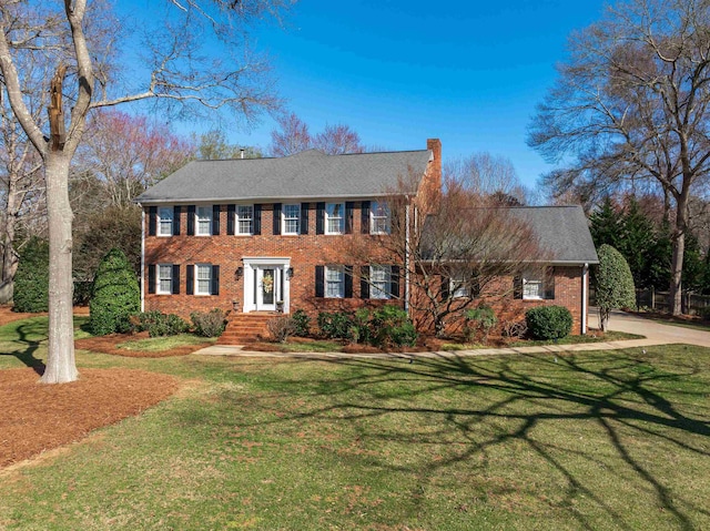 colonial inspired home featuring a shingled roof, a chimney, a front lawn, and brick siding