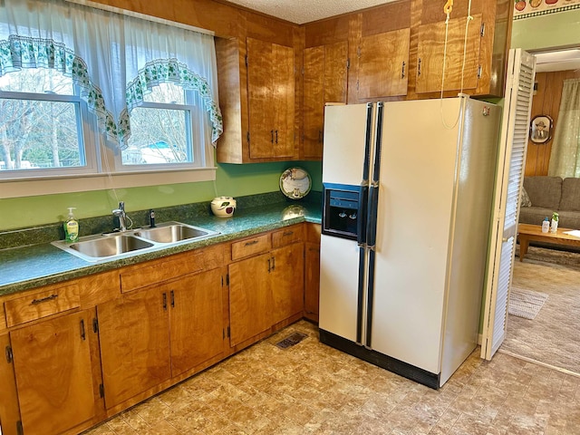 kitchen featuring white refrigerator with ice dispenser, brown cabinetry, dark countertops, and a sink