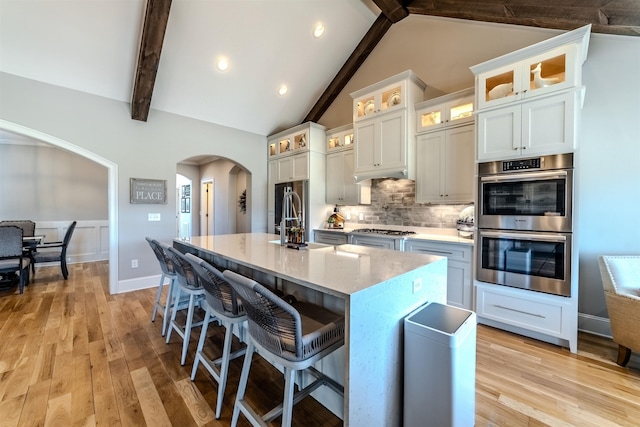 kitchen featuring arched walkways, vaulted ceiling with beams, a center island with sink, stainless steel appliances, and decorative backsplash