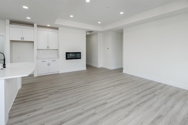 unfurnished living room with recessed lighting, a raised ceiling, a glass covered fireplace, light wood-type flooring, and baseboards