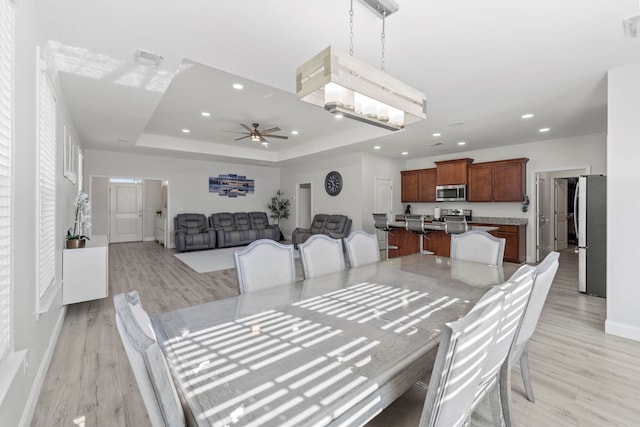 dining room with recessed lighting, ceiling fan with notable chandelier, baseboards, a tray ceiling, and light wood finished floors